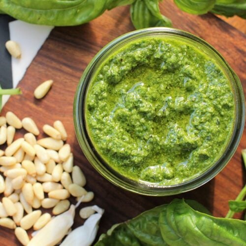 classic basil pesto, with pine nuts and garlic, looking down into the jar, with a pile of pine nuts beside it.