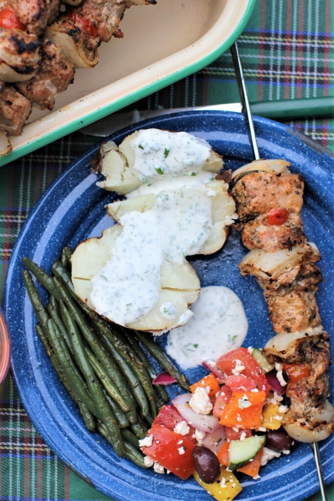 blue tin plate with souvlaki skewer, baked potato, grilled beans and Greek salad