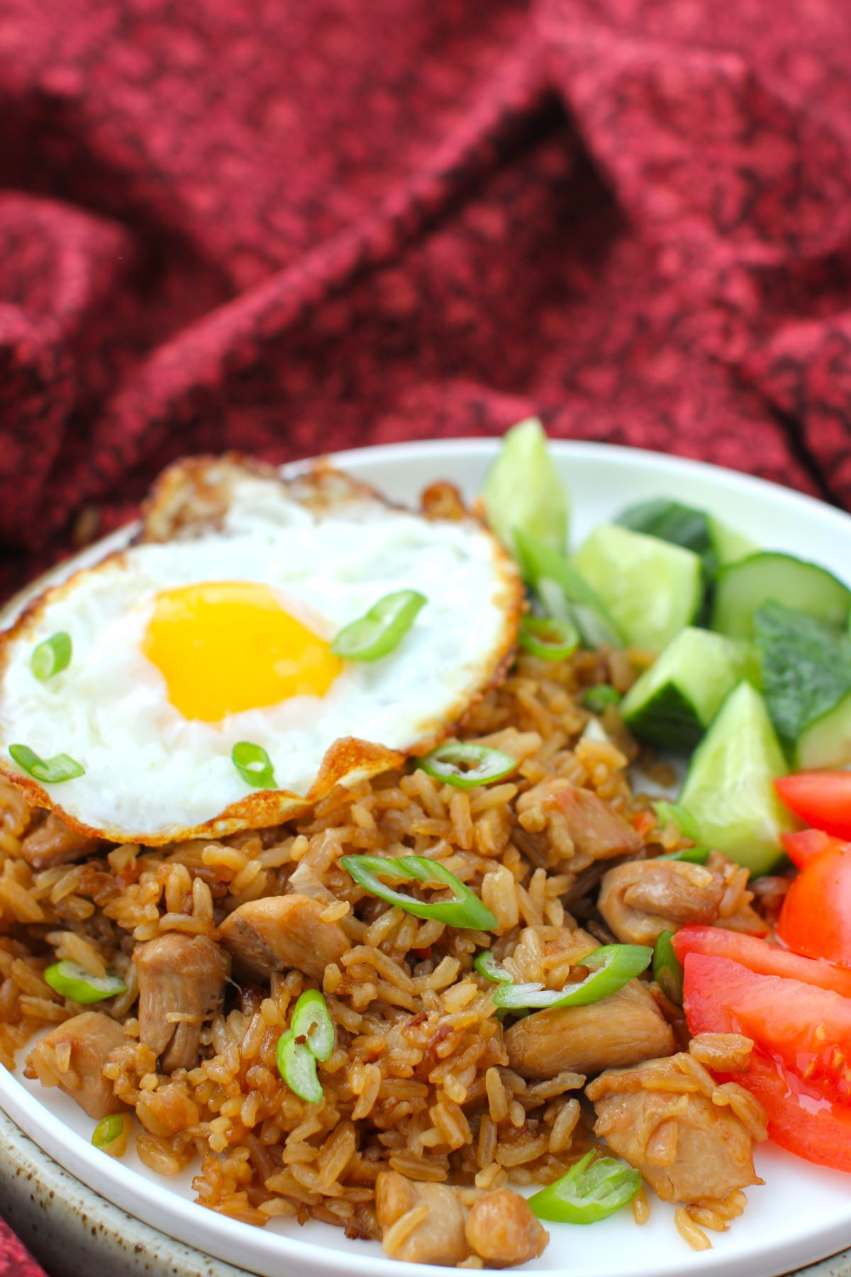 plate of Indonesian fried rice, with fried egg on top, tomatoes and cucumbers, against a burgundy cloth.