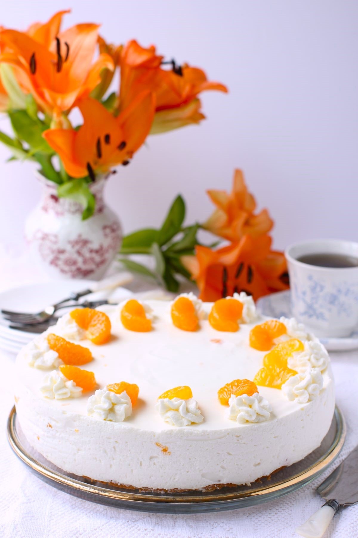 A Mandarin Orange Cream Torte on the table with a vase of orange lilies behind it.