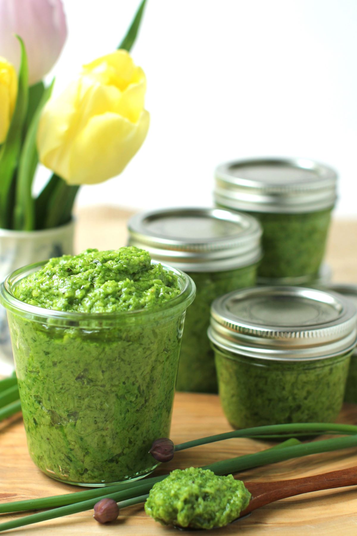 A jar of chive pesto on a table, with closed jars of it behind, and a vase with spring tulips.