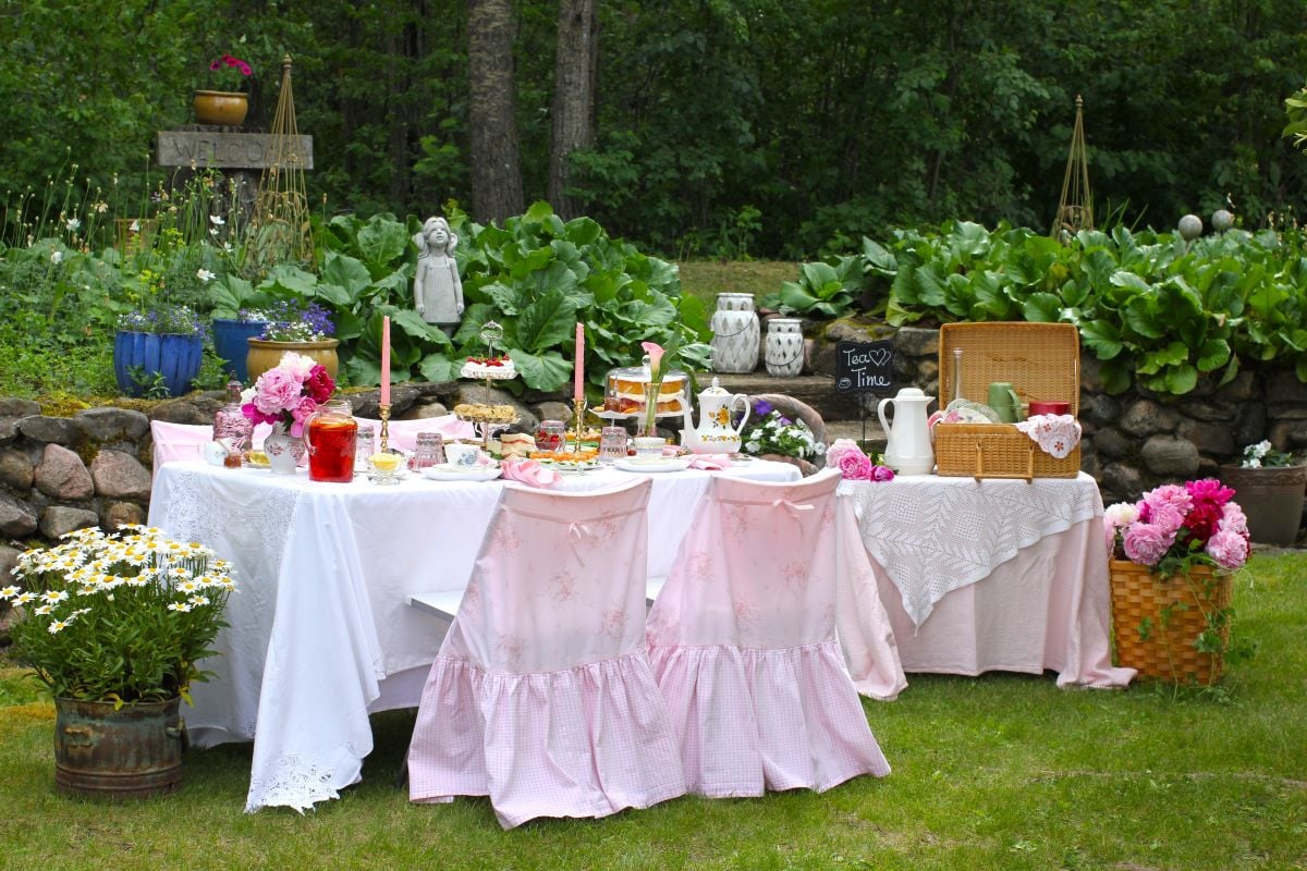 The table set outdoors for a lovely, pink-inspired afternoon tea picnic outside. Table set with three-tiered stand of treats and a cake stand with a gluten free Victoria sponge cake, by the rock wall.