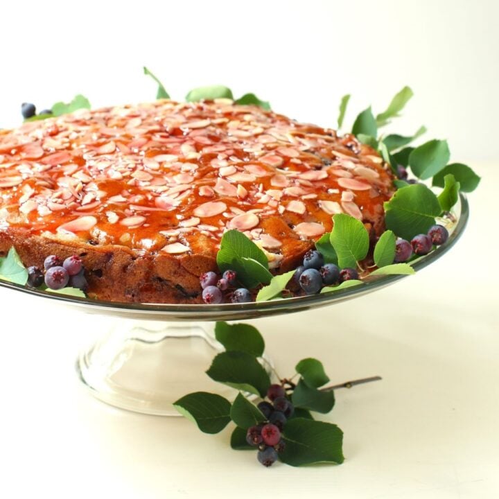 The Saskatoon Drizzle Cake on a glass cake stand, surrounded with sprigs of fresh saskatoon berries.