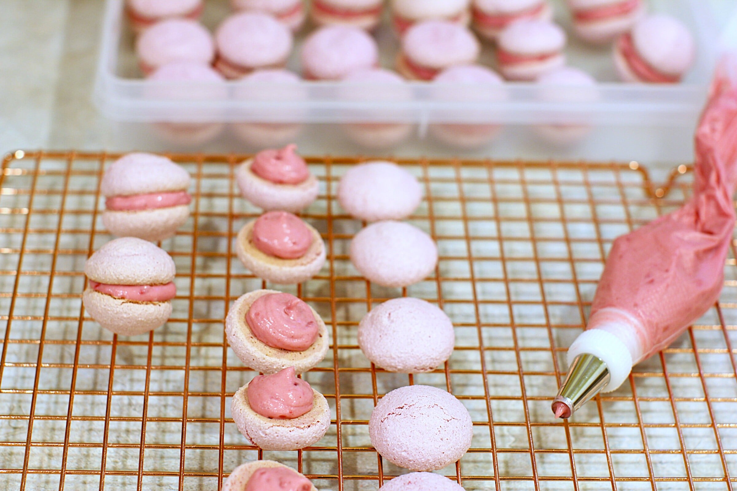Cooling rack with raspberry meringue shells, some with filling piped on, and piping bag beside.