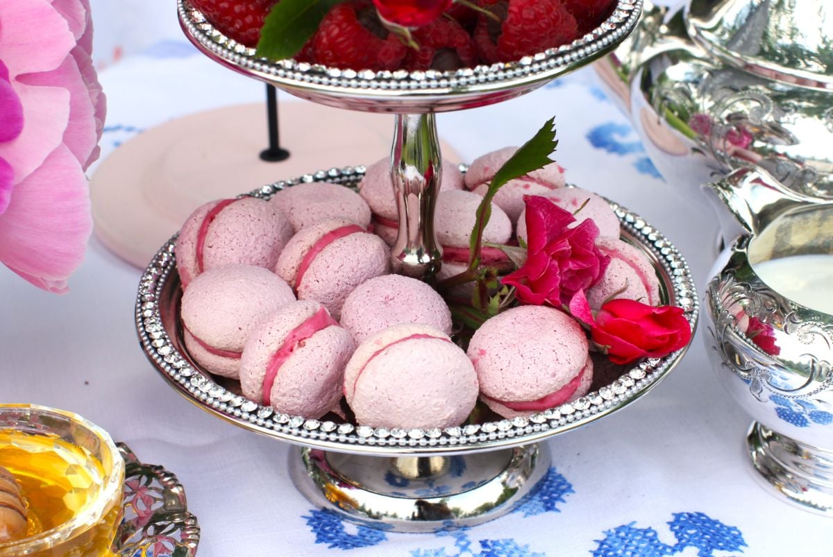A 2-tiered silver cake stand filled with pink Raspberry Meringue Kisses on the bottom and chocolate ganache-filled raspberries on top.