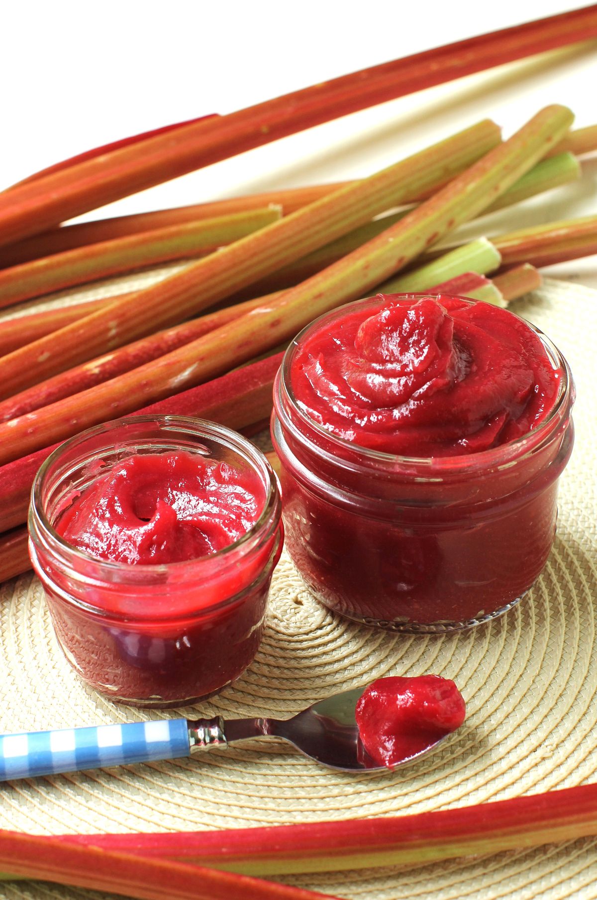 Two jars of rhubarb butter, one large, one small, with the rhubarb butter swirled up high in it. Stalks of rhubarb behind and a spoonful of rhubarb butter in front.