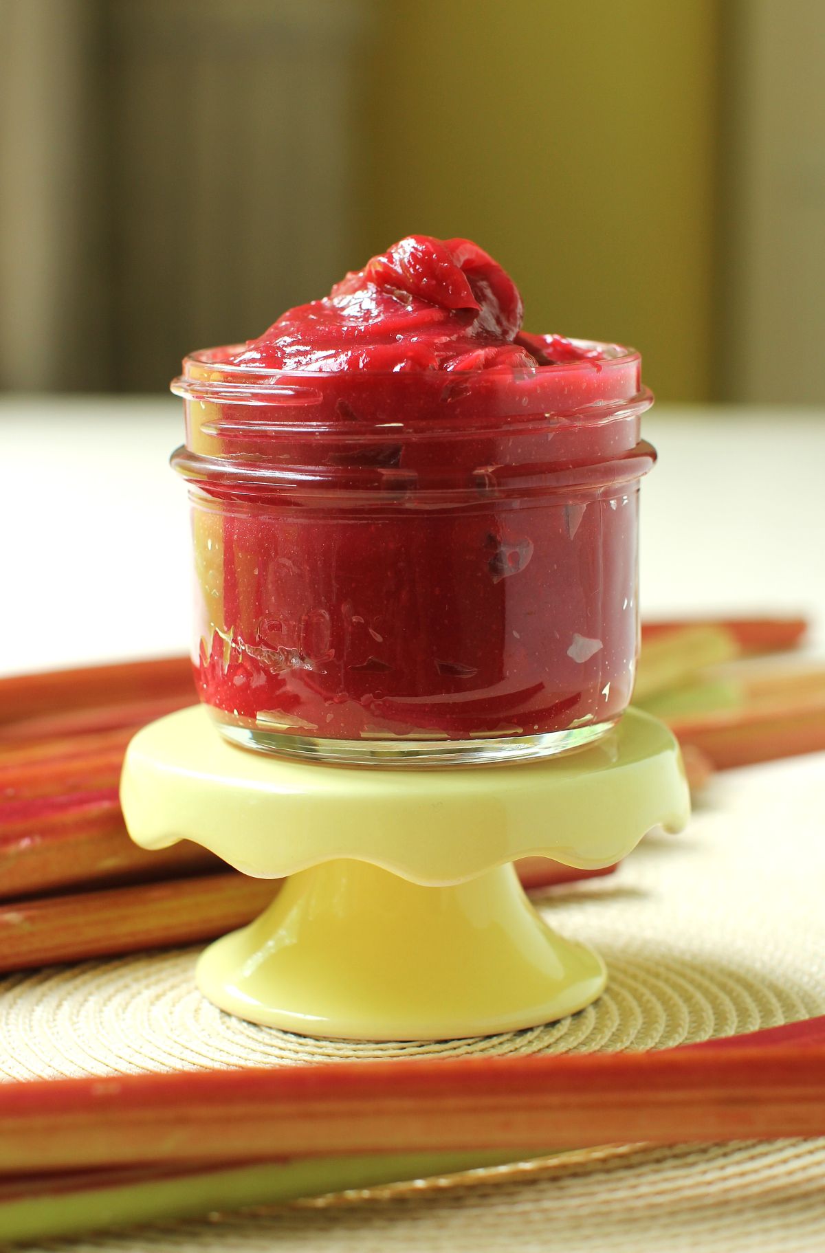 A jar piled high with a mound of rhubarb butter, sitting on a small footed yellow stand, with rhubarb stalks around it. 