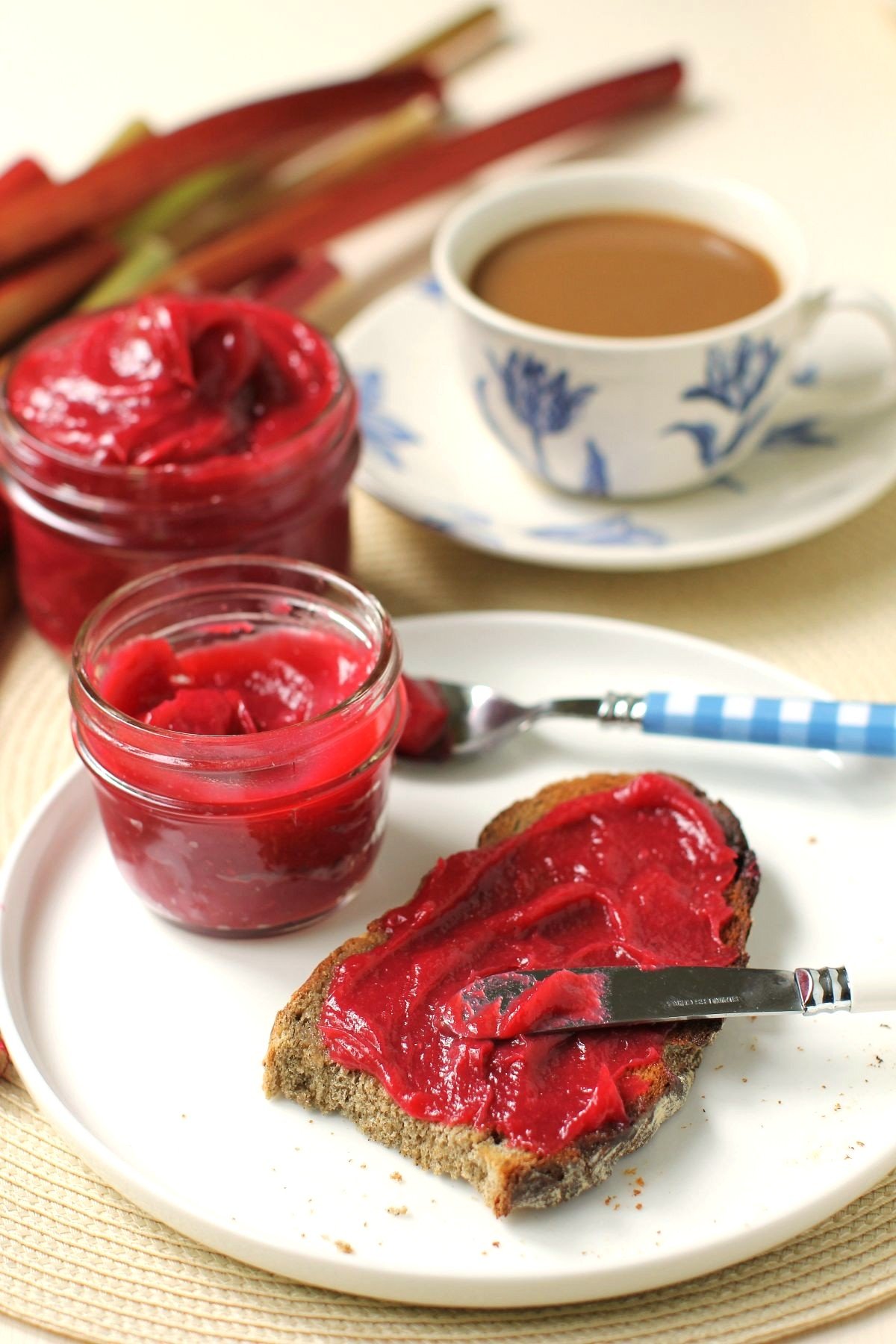 Rhubarb butter slathered on toast with a cup of coffee behind it and more rhubarb stalks.
