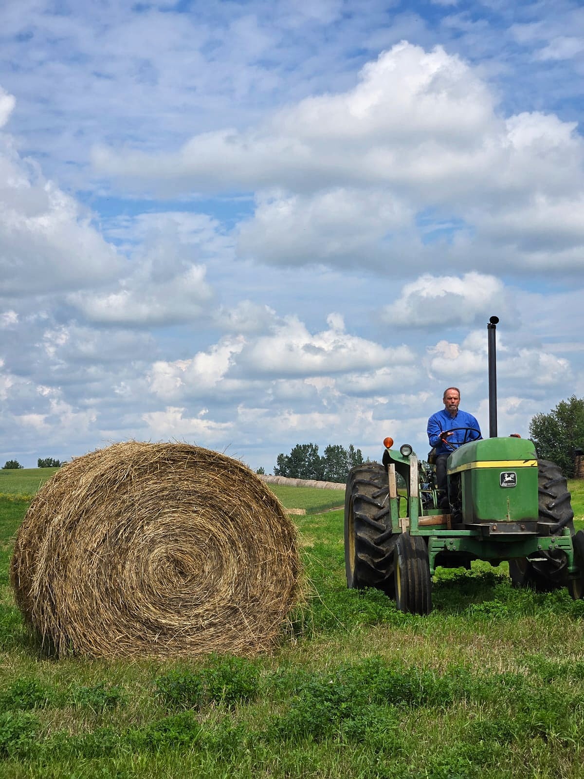 Raymond driving the old green tractor beside a big round bale of hay.
