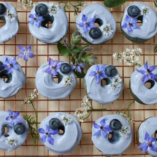 Square thumbnail photo. Mini blue donuts laid out in a grid pattern on a copper cooling rack, with borage flowers and cilantro flowers tucked in betwen them.
