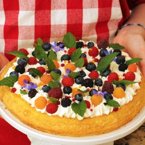 Thumbnail photo of Amelia holding the fruit & flower almond cake. Red & white checked apron in background.