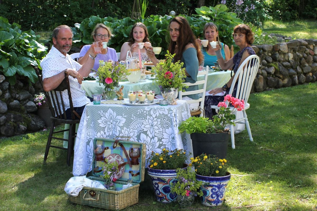 6 people sitting around the laden afternoon tea table, sipping on tea cups.