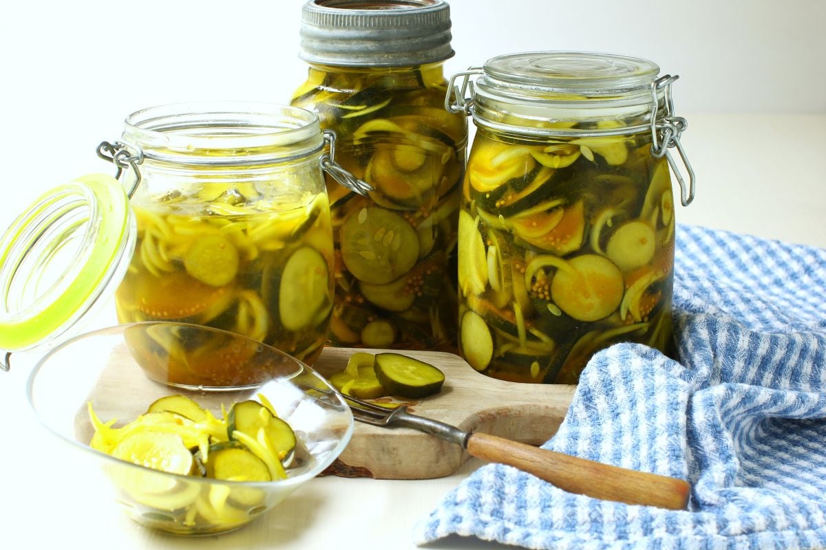 Horizontal photo of 3 jars refrigerator bread & butter pickles with some in a bowl, blue & white tea towel beside.