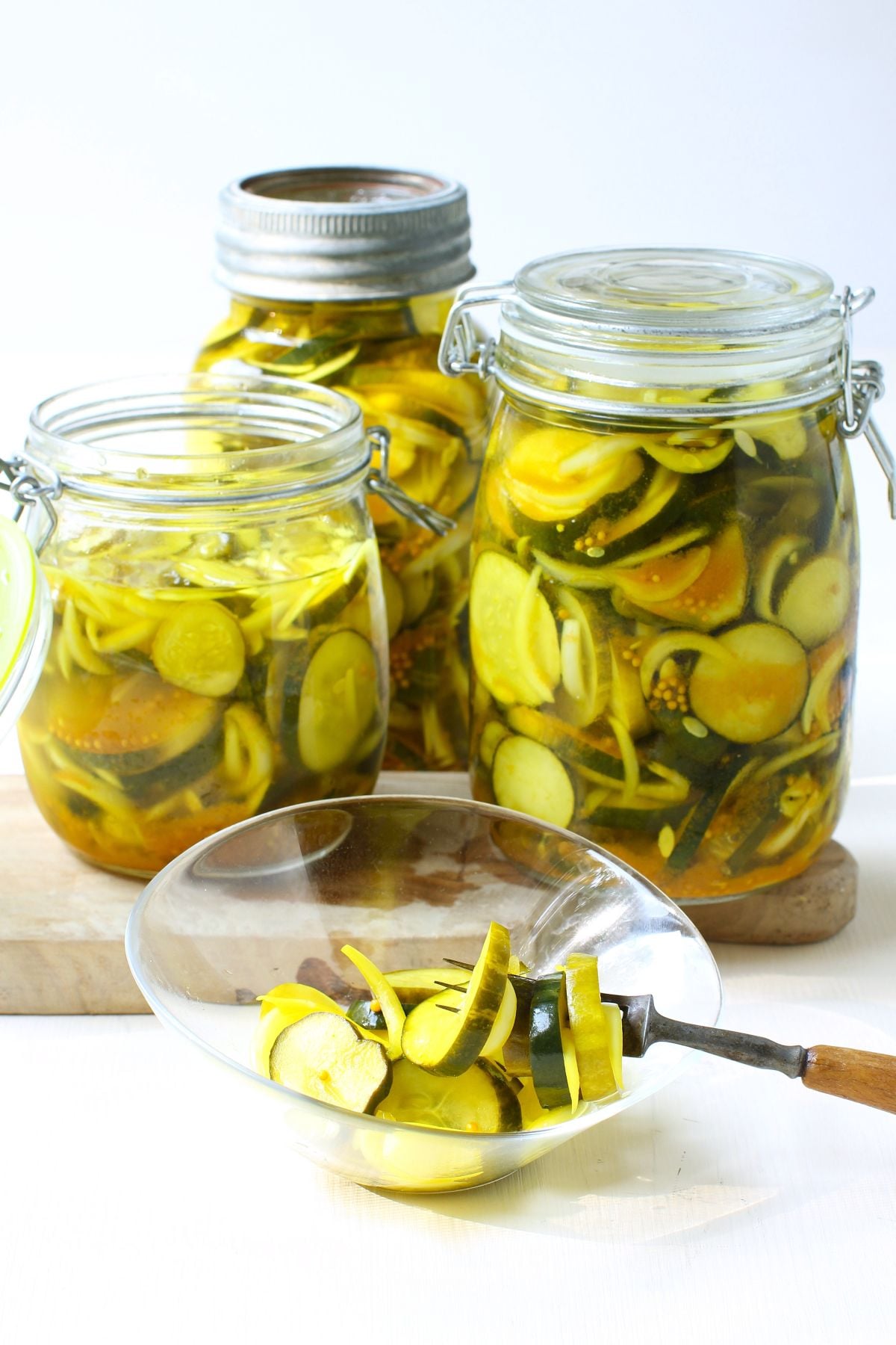 3 jars of refrigerator bread & butter pickles, with some in a small bowl in front and a fork holding some pickle slices, too.