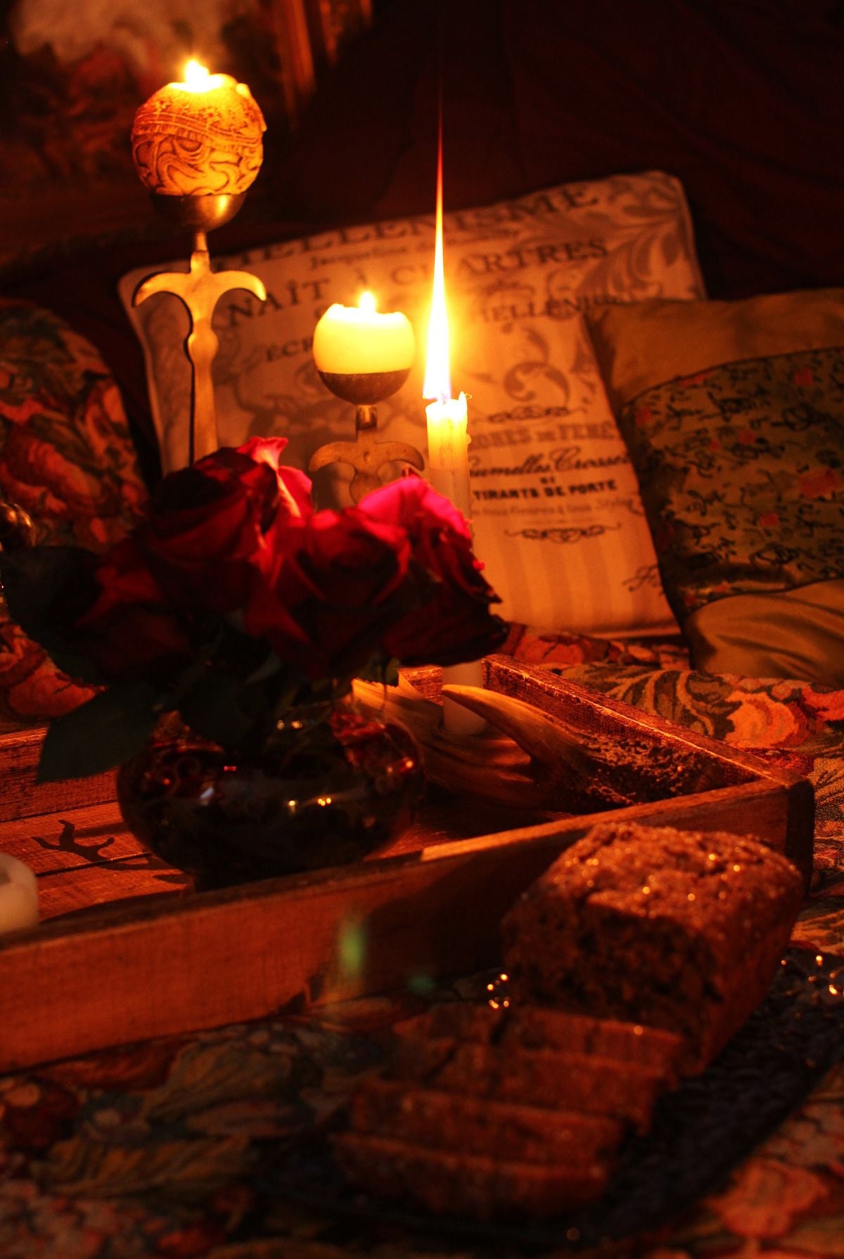 Round candle and short candle glowing in the dark on a wooden tray, with bouquet of dark roses and bara brith cake in front.