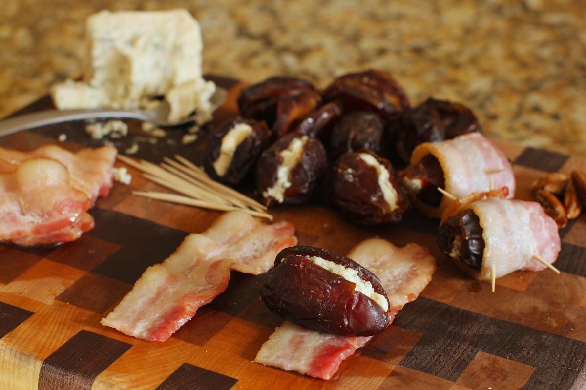 Checkered cutting board with stuffed dates, bacon, and cheese, and a stuffed date laying on a piece of bacon, ready to roll up.