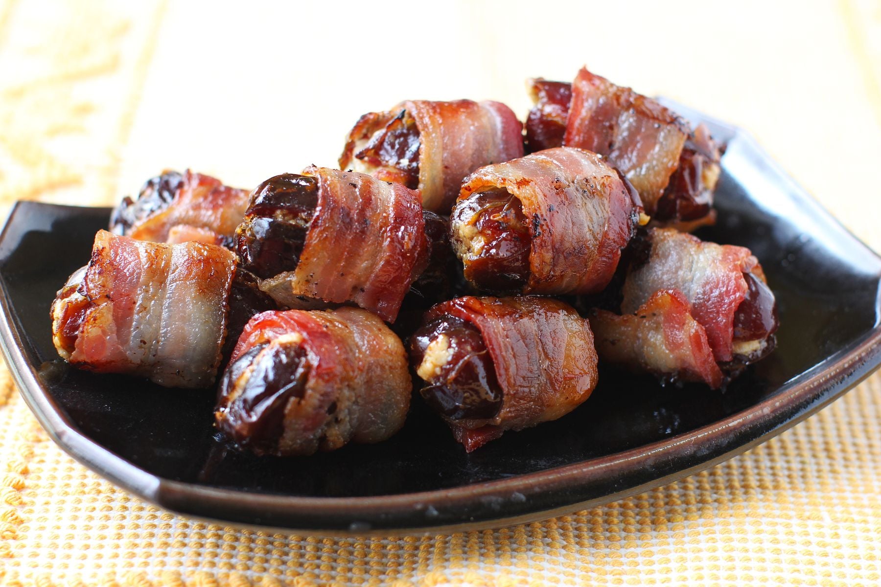 Black plate with a heap of bacon wrapped dates on it, on yellow mat.