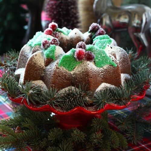 square thumbnail photo of gingerbread bundt cake, decorated and on a red pedestal cake stand with spruce boughs.