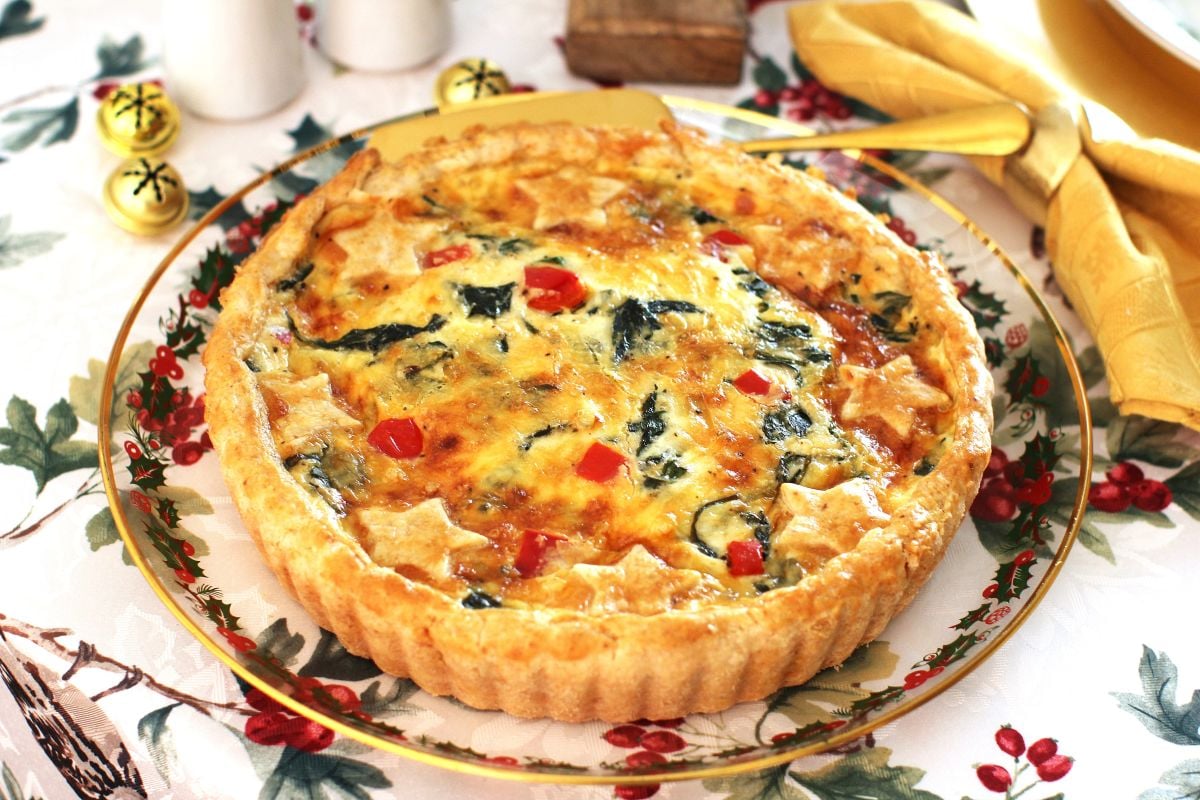 Golden and beautifully baked Christmas quiche on a holiday platter, set on top of a Christmas tablecloth.