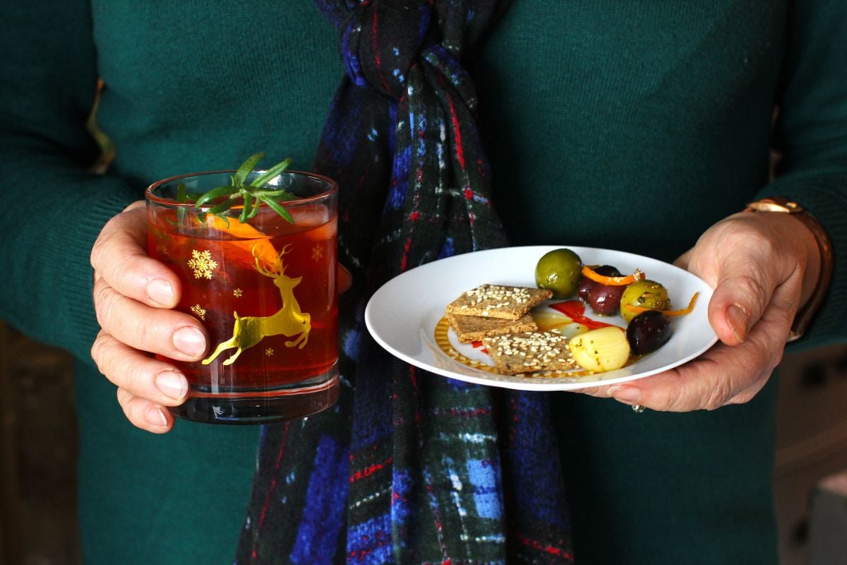 A woman in green dress holding a cocktail in one hand and plate of olives and crackers in the other.