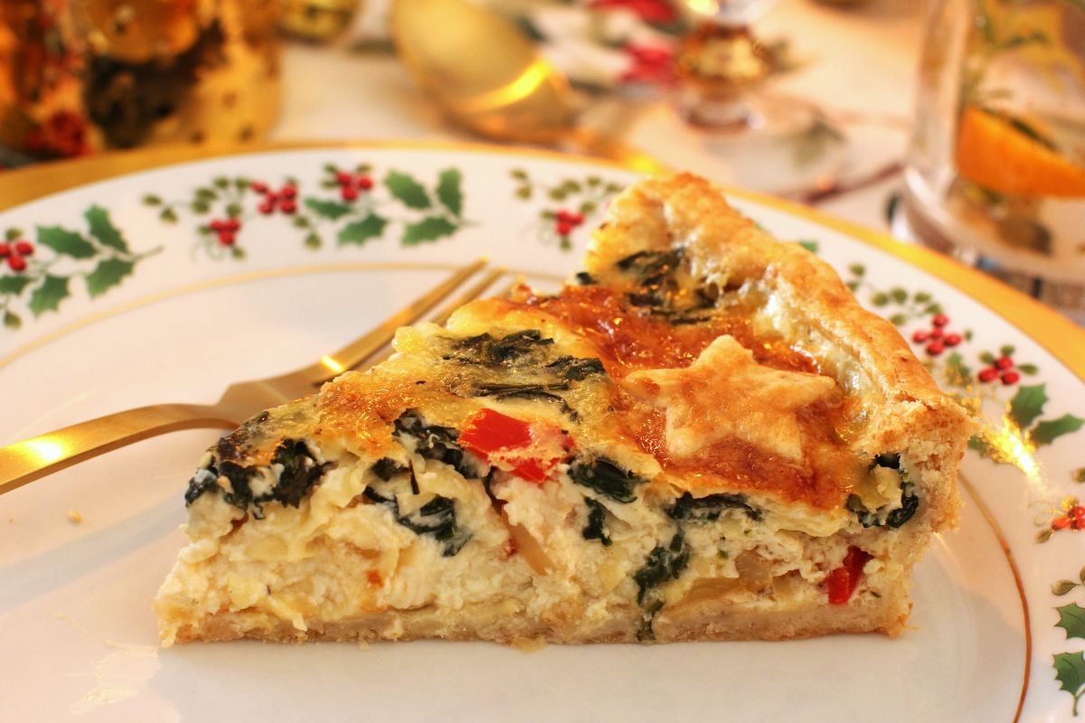 A slice of Christmas quiche on a holly-rimmed white plate with a gold fork beside it.