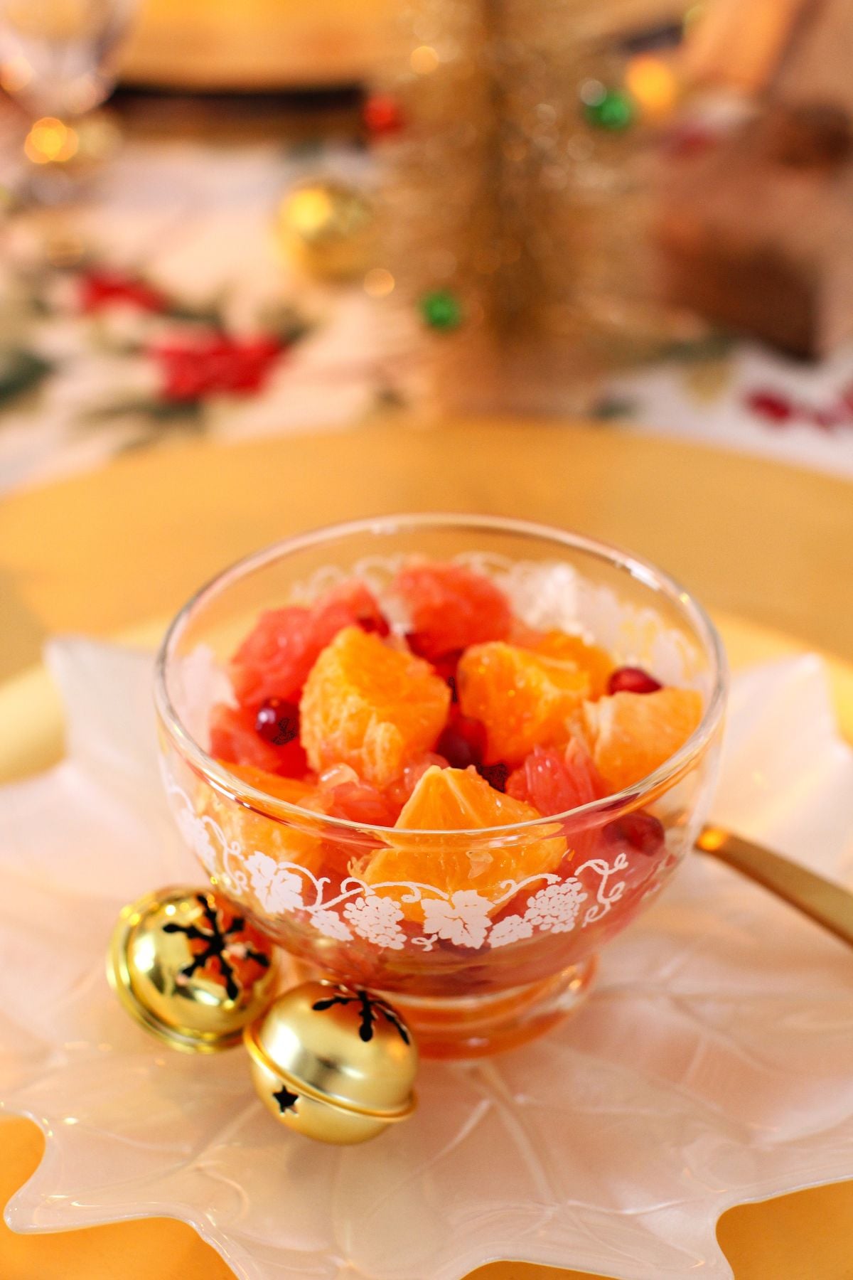 A small glass fruit nappy with citrus salad on white plate, with two golden jingle bells beside it.