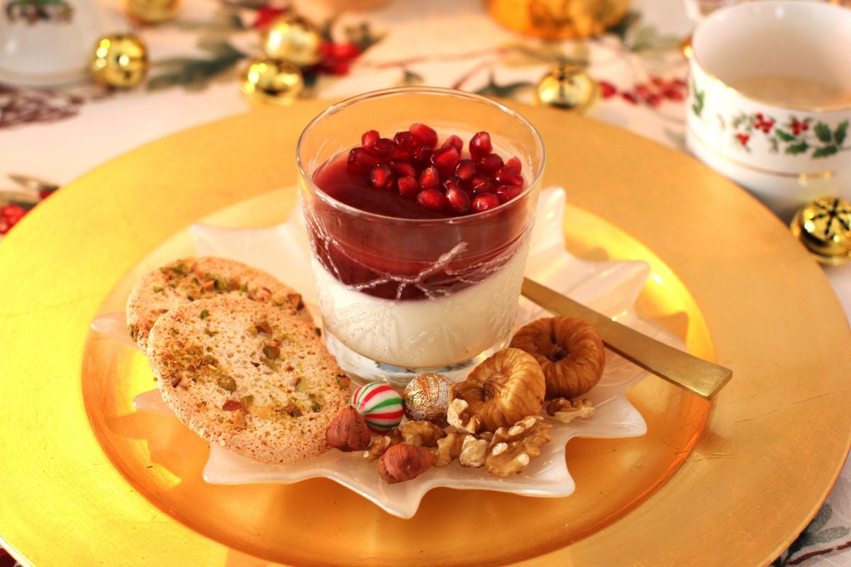 Yogurt pannacotta with pomegranate jelly in a glass on a gold plate, two almond dacquoise biscuits beside, and dried figs, nuts, and chocolates.