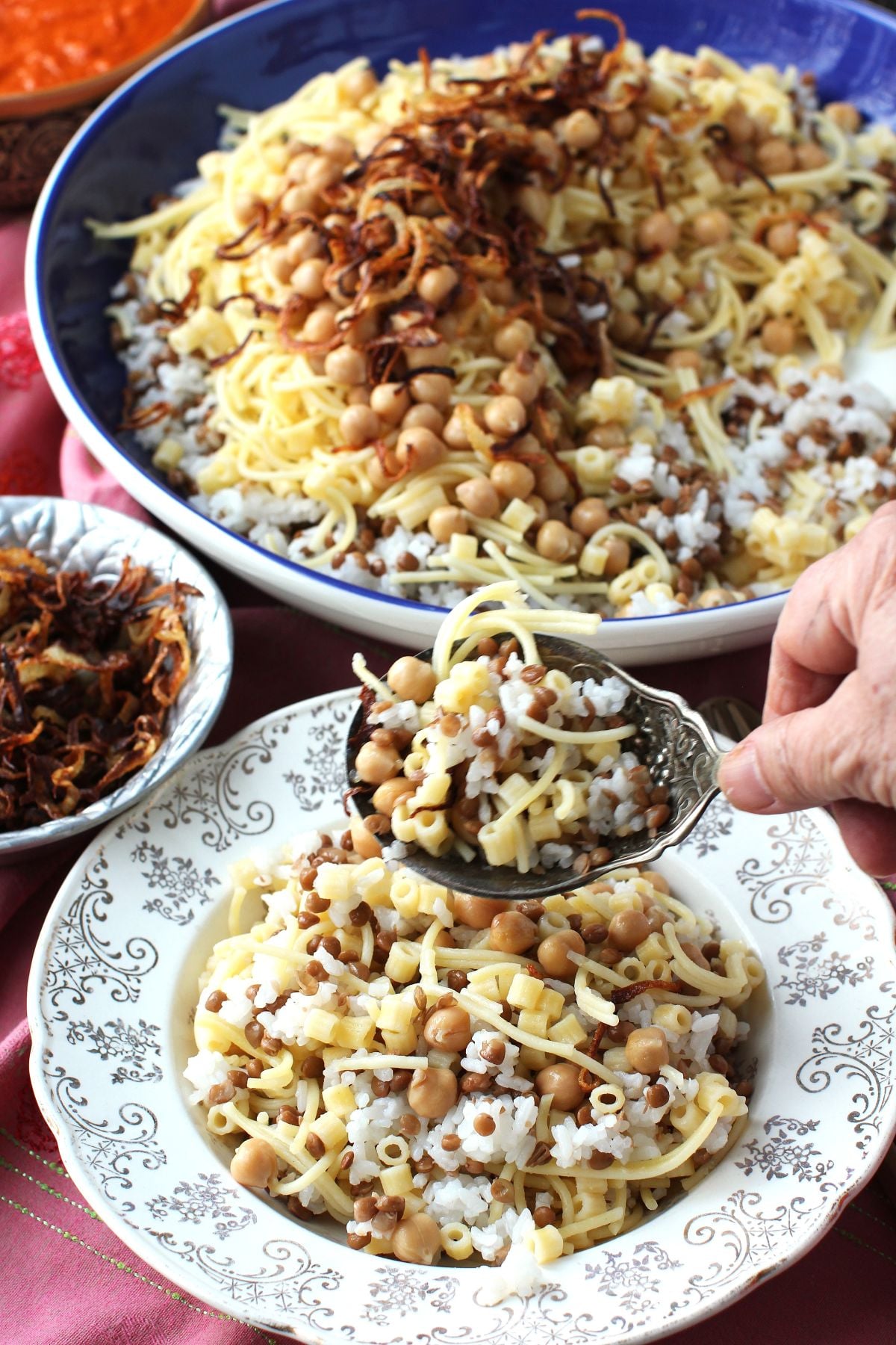 Adding a scoop of koshari main ingredients to an individual bowl from the larger blue serving bowl.