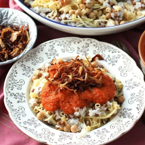 Square photo of plate of koshari with tomato sauce and fried onions on top. Thumbnail photo.