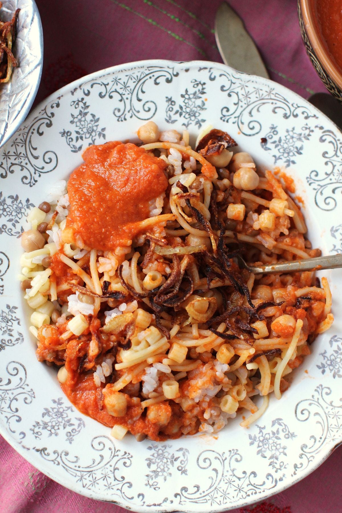Top-down view of a plate of koshari, stirred up, with a dollop of tomato sauce and a few crispy fried onions on top.
