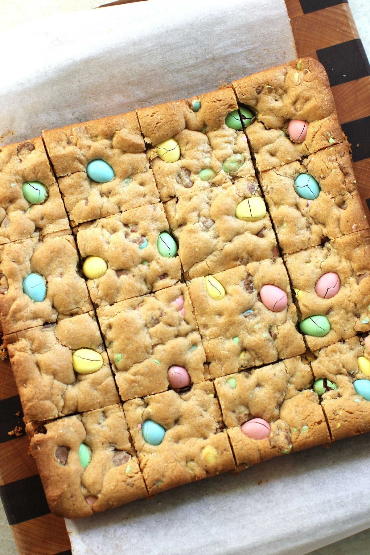 Slab of Mini Egg Cookie Bars on parchment paper on cutting board, just cut into 16 squares.