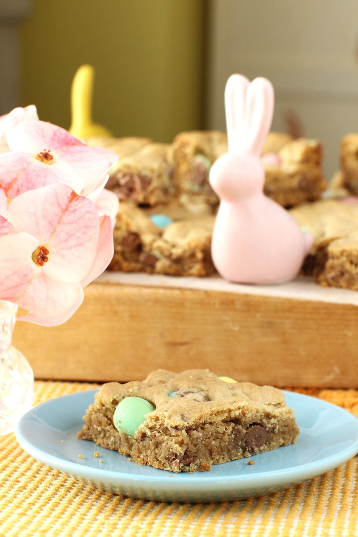 A Mini Egg Cookie Bar on a light blue small plate, with the rest on a cutting board behind it, and small porcelain bunnies.