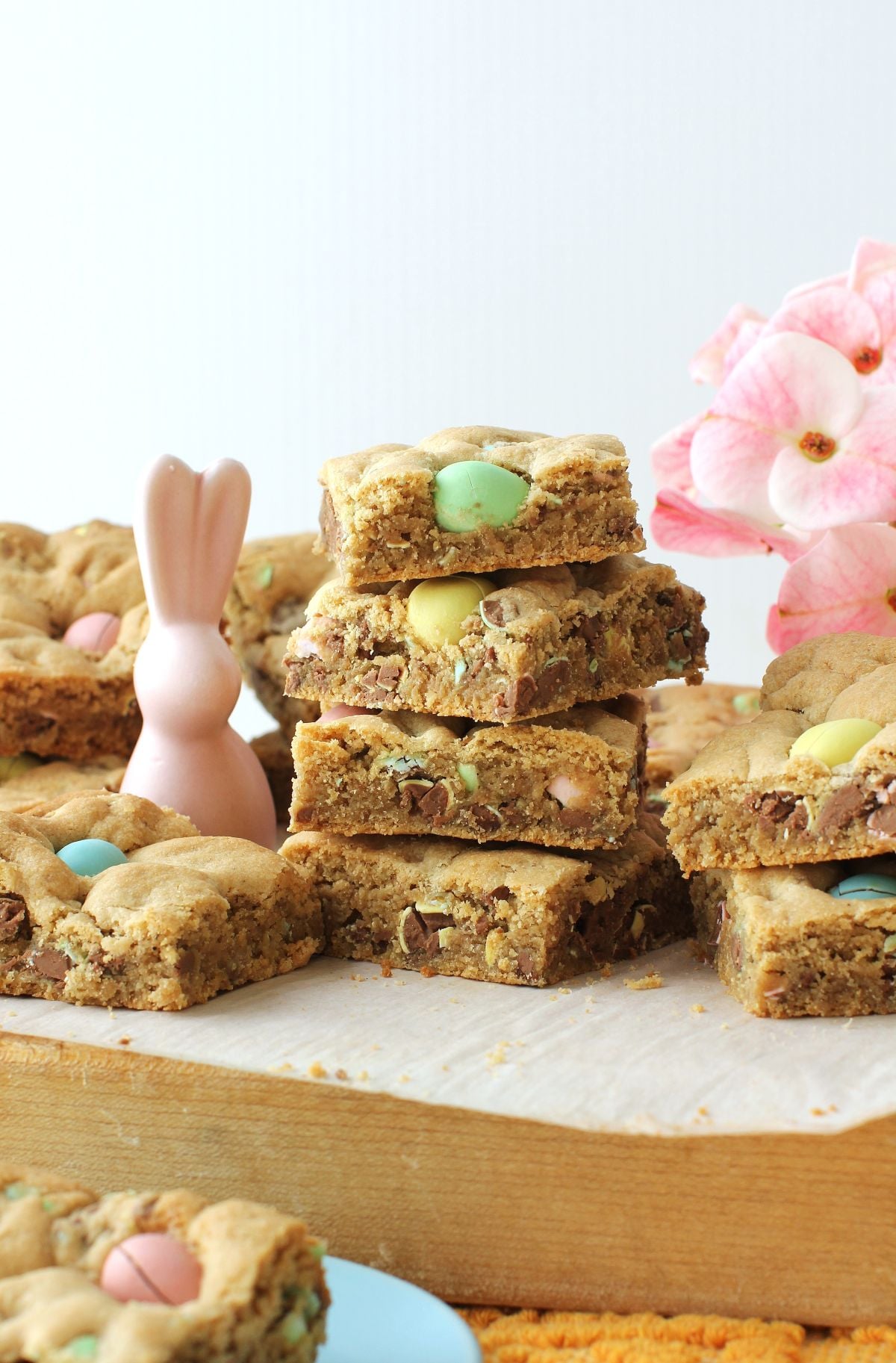 Stack of mini egg cookie bars on a cutting board, with pink flowers and pink porcelain bunny.
