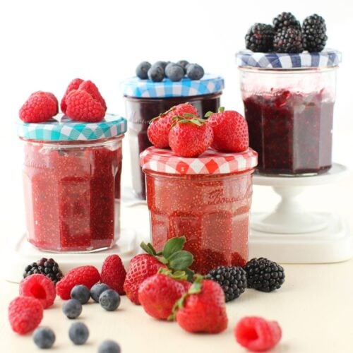 4 jars of different chia berry jams with colourful lids. Corresponding berries on top of each lid and scattering of berries in front.