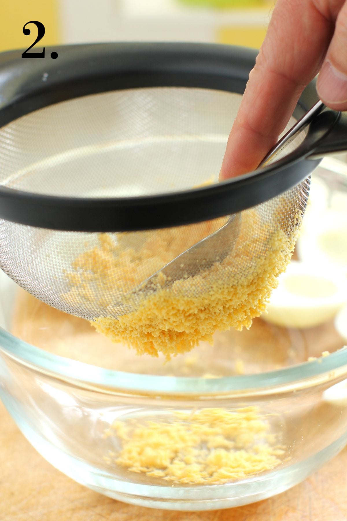 Pressing down on the cooked yolks with the back of a spoon to push them through a large meshed sieve into a clear glass bowl below.