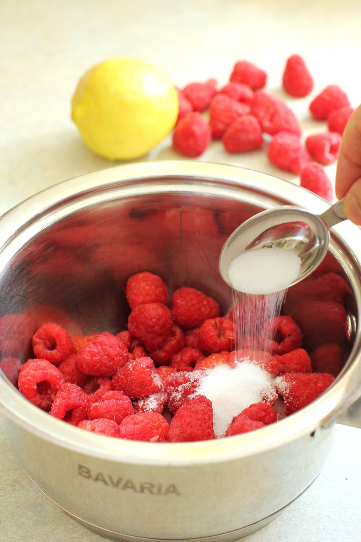 Adding a tablespoonful of sugar to raspberries in a stainless steel pot.
