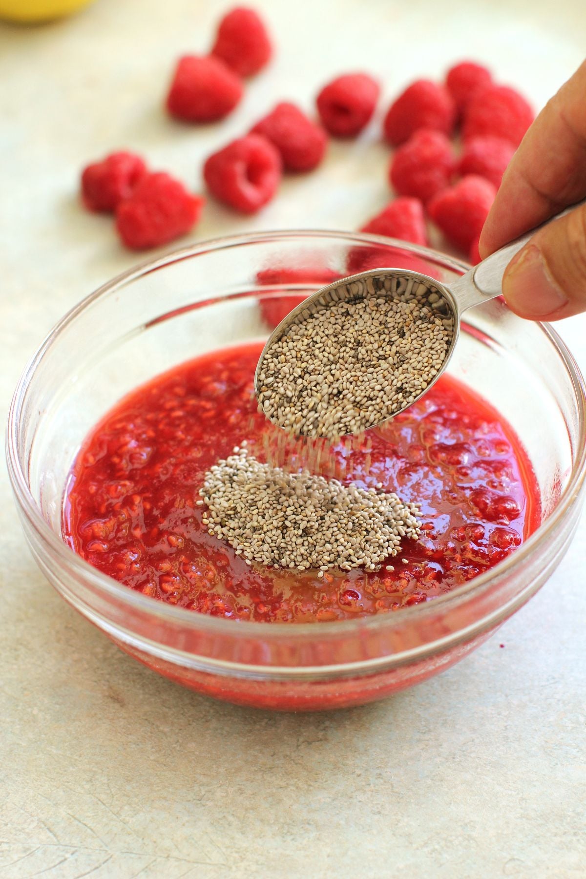 Spooning chia seeds into the raspberry jam in a small clear glass bowl.