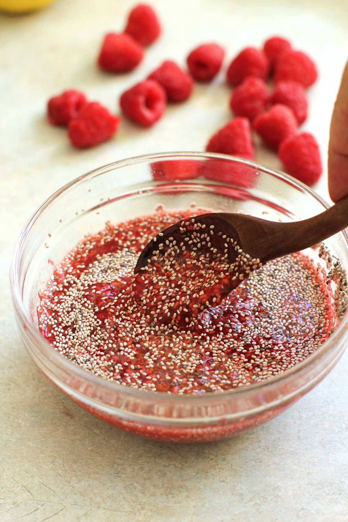 Stirring chia seeds into raspberry jam in a small clear glass bowl with a wooden spoon.