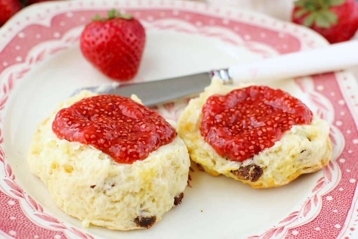 A close-up photo of a scone split and spread with strawberry chia jam on a pink and white plate, white handled spreading knife, one strawberry on the plate.