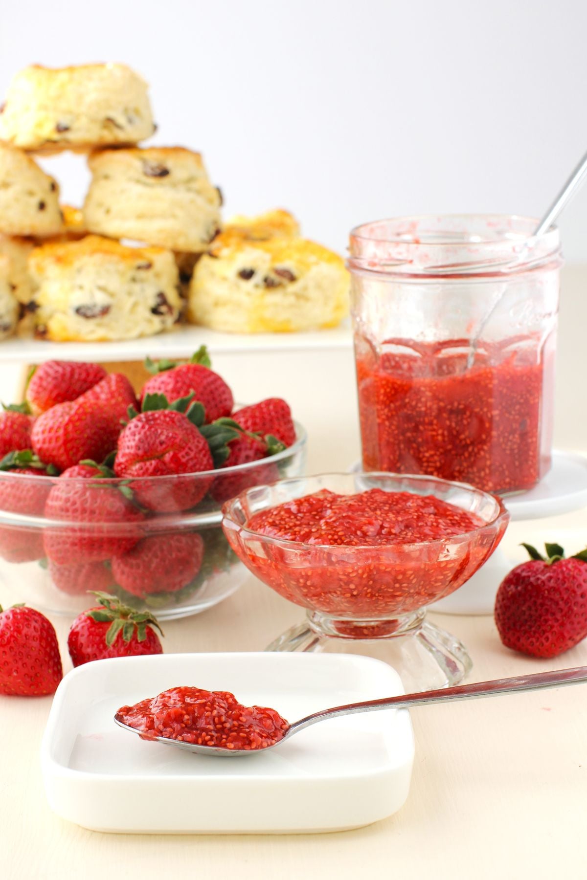 Spoonful of strawberry chia jam on a small white plate, bowl of strawberries, footed glass bowl of jam, jar of jam, and raised plate of stacked scones behind, on white background.