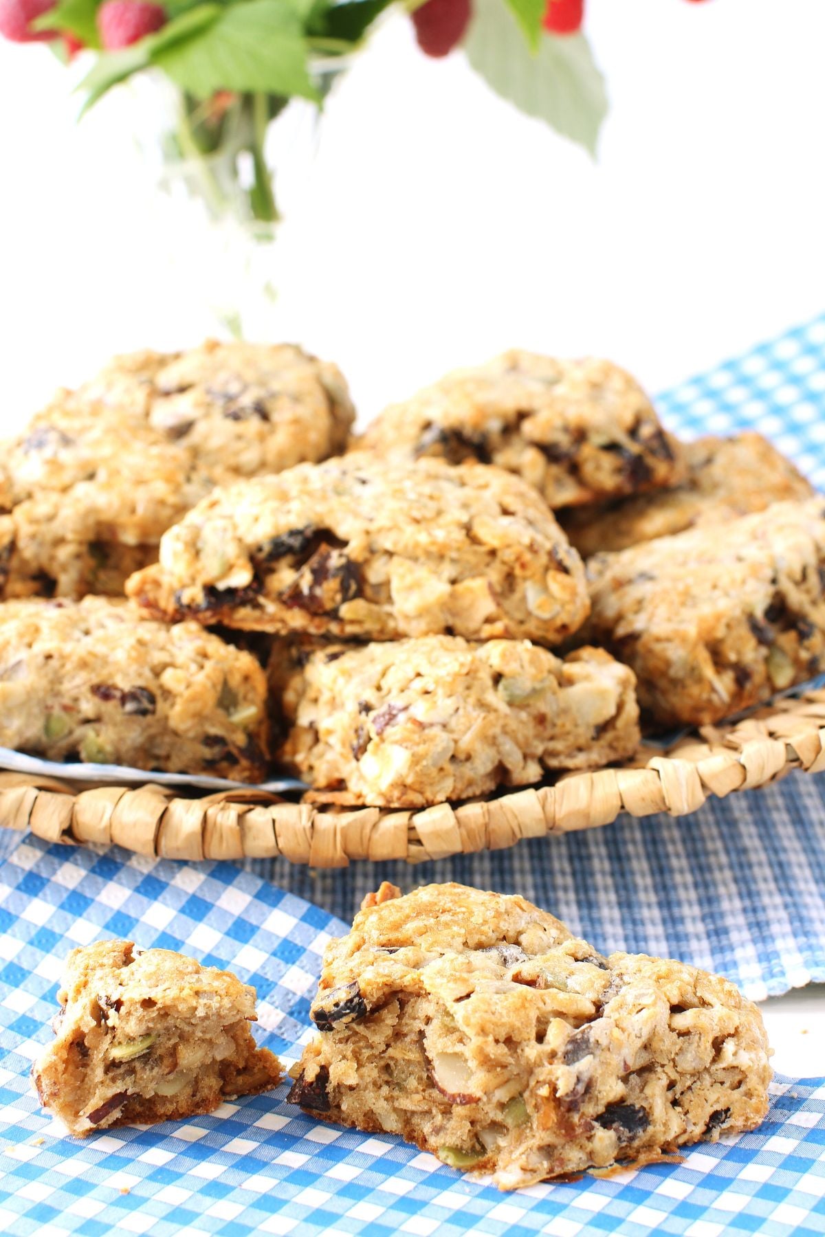 plate of granola scones in background, and one laying on a blue and white checked napkin in front, a piece broken off and ready to eat. a cluster of raspberries and leaves in top left of photo.