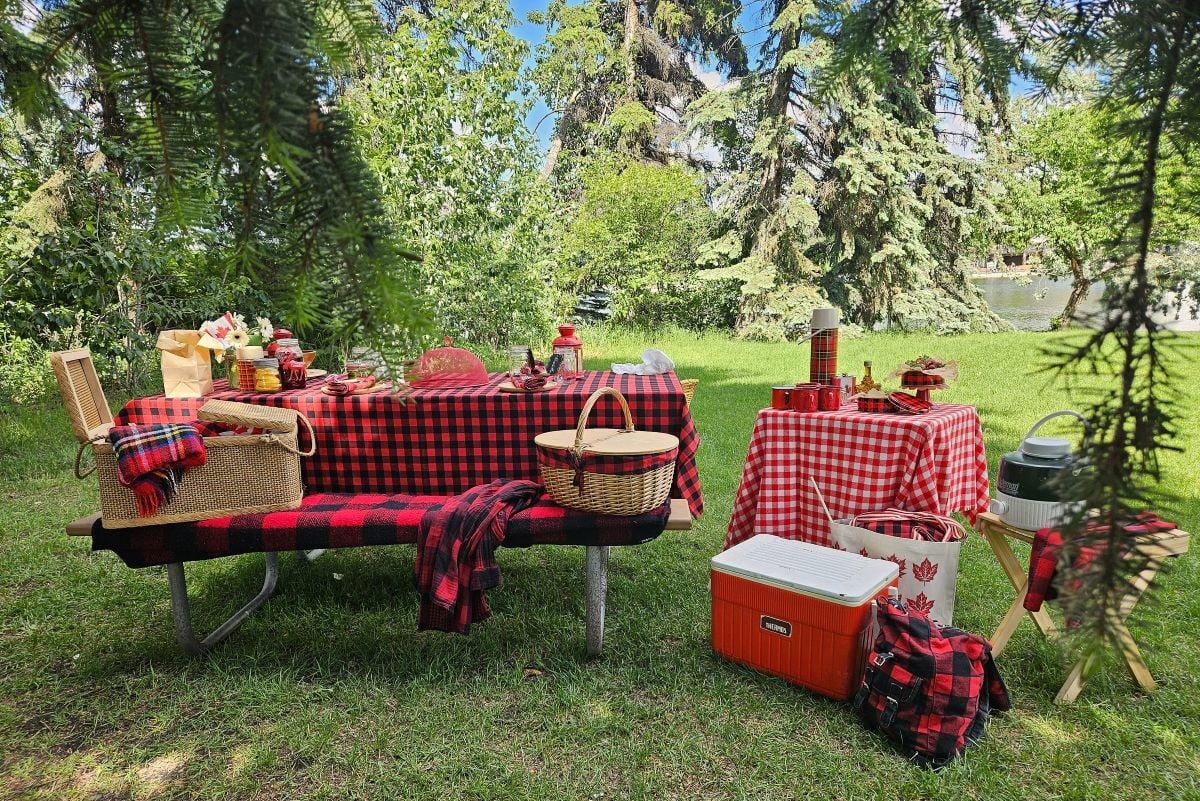 Picnic table set in the grass and trees, covered with a red and black checked tablecloth and dishes, wicker picnic baskets, and a side table with red and white checked cloth, red and white cooler in front, and red and black backpack.