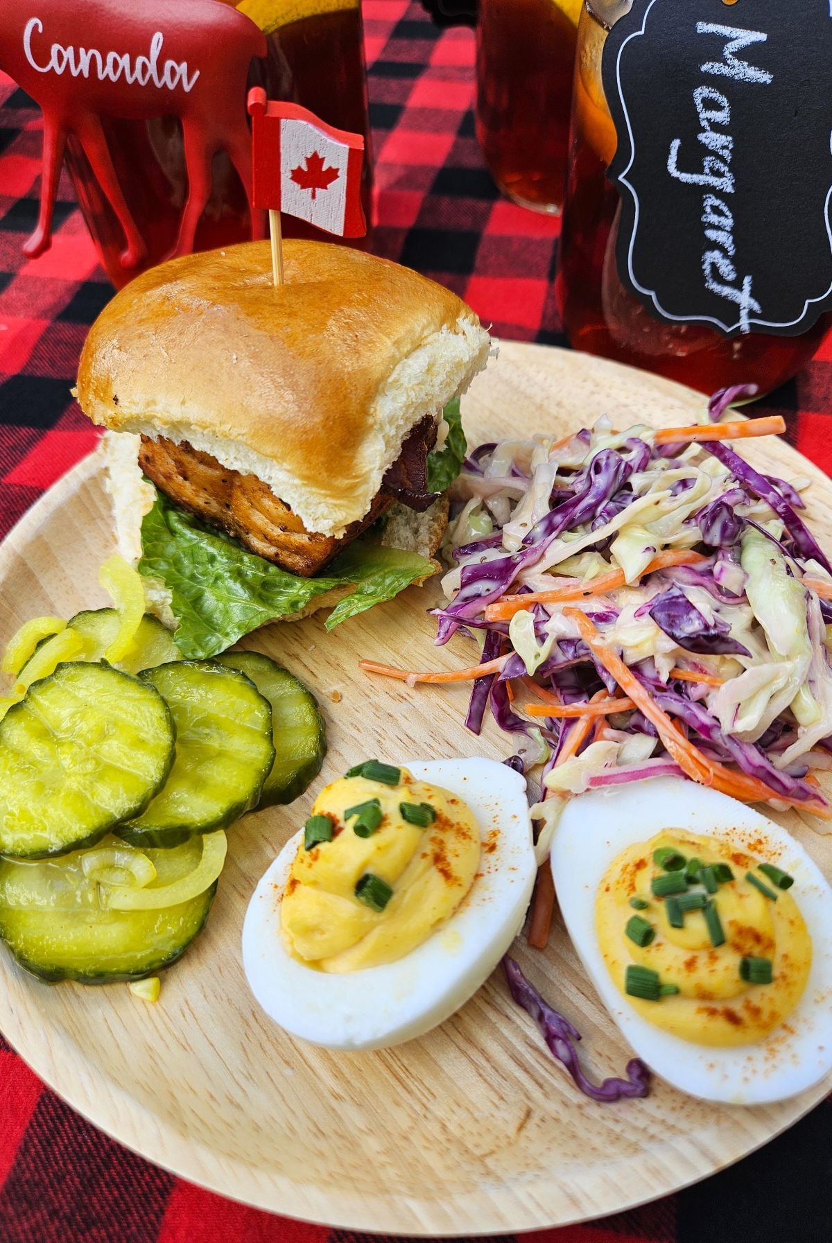 Small wooden plate full of Canadiana picnic food - a salmon slider bun, coleslaw, bread & butter pickles, and 2 deviled egg halves.