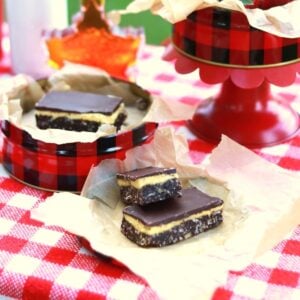 Square thumbprint photo of Nanaimo Bars on crumpled brown parchment paper on red and white checked tablecloth.