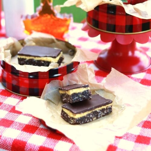 Square thumbprint photo of Nanaimo Bars on crumpled brown parchment paper on red and white checked tablecloth.