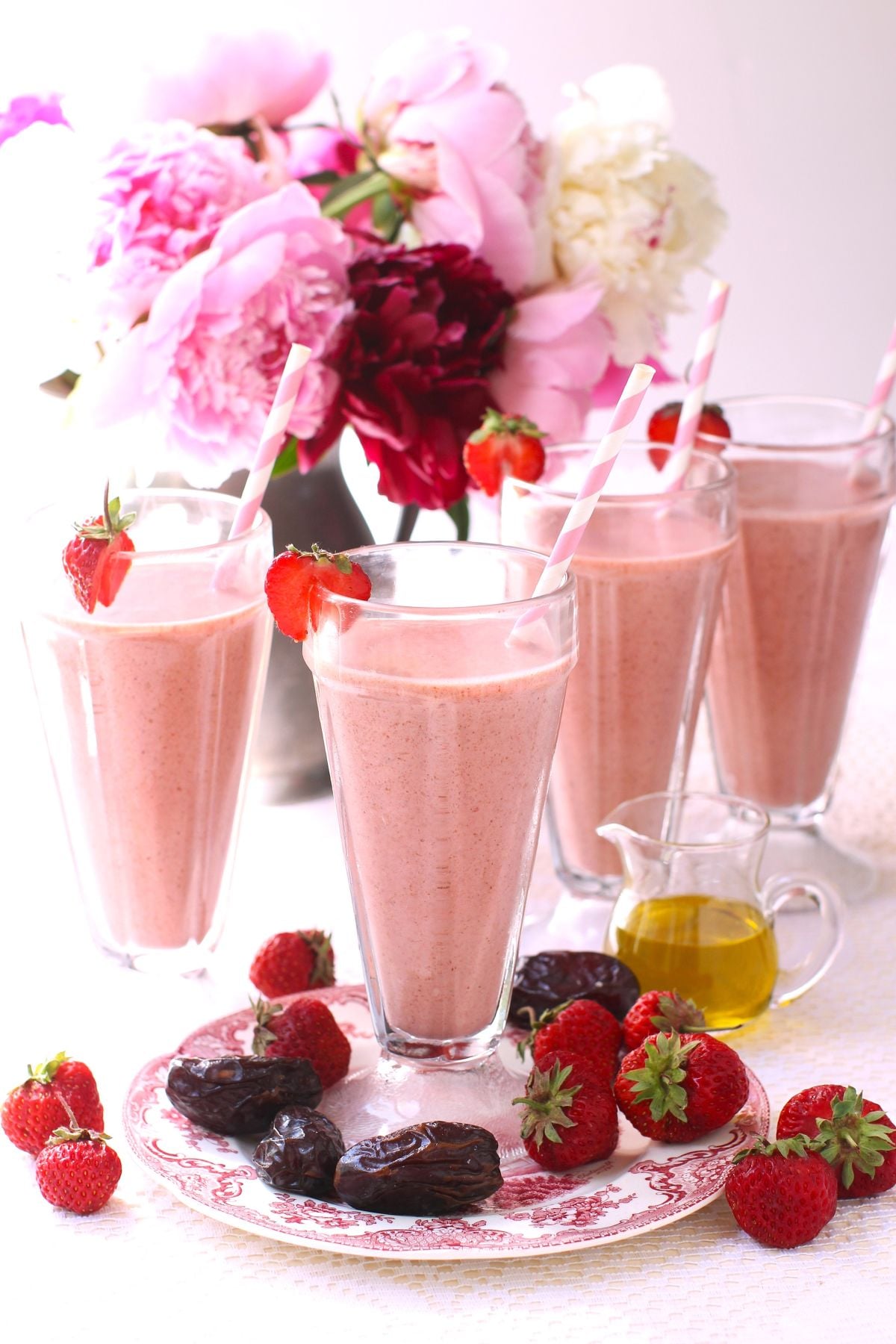 4 tall stemmed milkshake glasses of pink strawberry date smoothies, with dates and strawberries on the table and a pitcher of pink peonies behind.