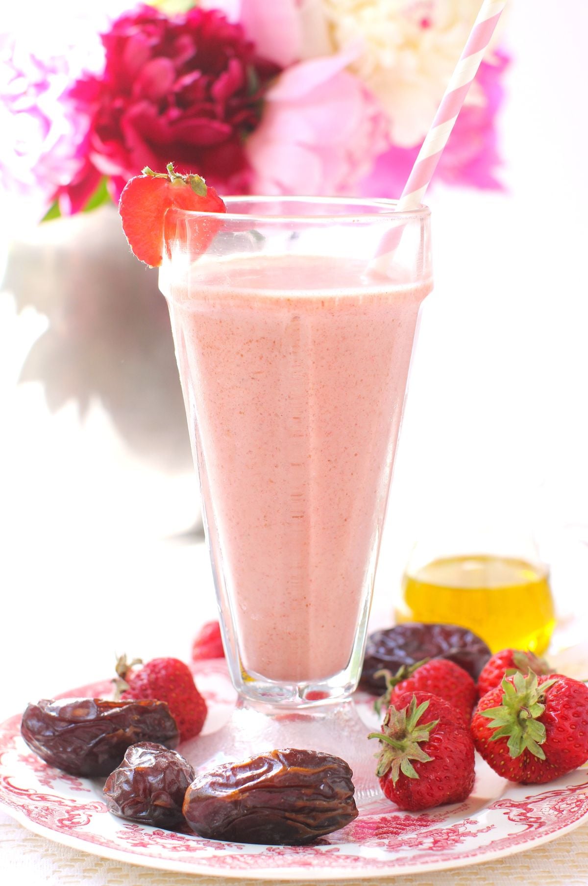 one tall glass of strawberry date smoothie on a pink plate with some dates and strawberries on it, pink flowers in background.