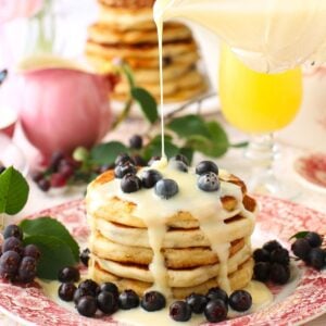 Square thumbnail photo of stack of saskatoon pancakes with berries on top and around, and a drizzle of sweetened condensed milk pouring down on them.