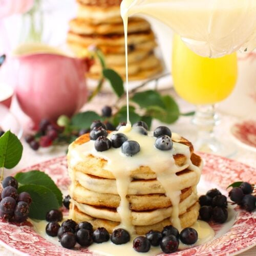 Square thumbnail photo of stack of saskatoon pancakes with berries on top and around, and a drizzle of sweetened condensed milk pouring down on them.