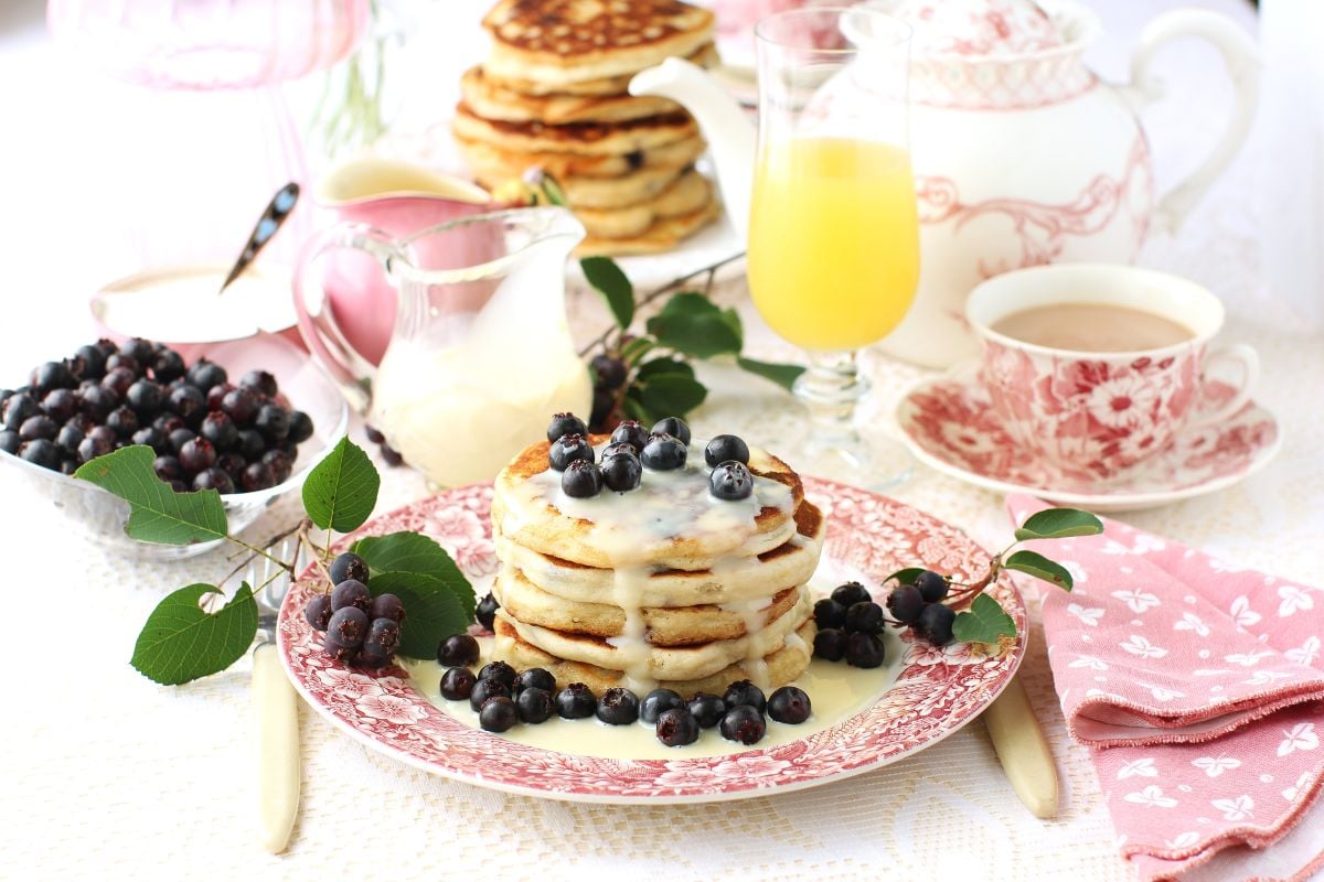 Breakfast table set, plate of saskatoon pancakes drizzled with condensed milk, glass of orange juice, pink dishes and napkin.